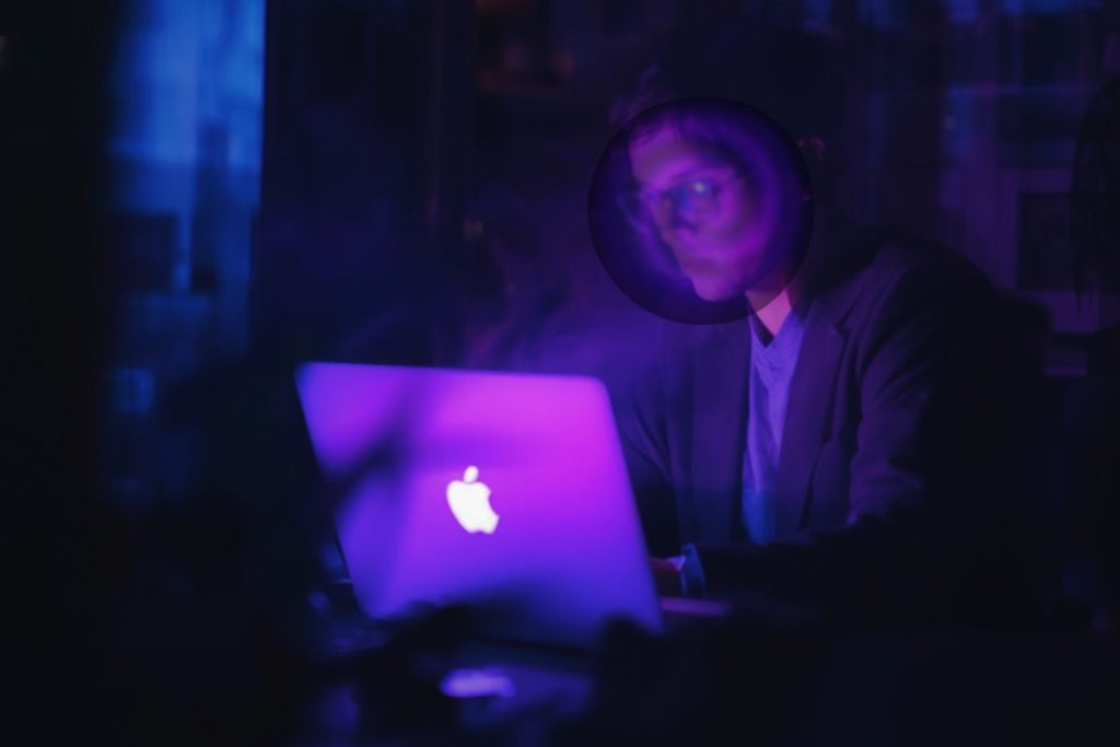 Man illuminated by a glowing laptop as his identity begins to blur into purple light and smoke