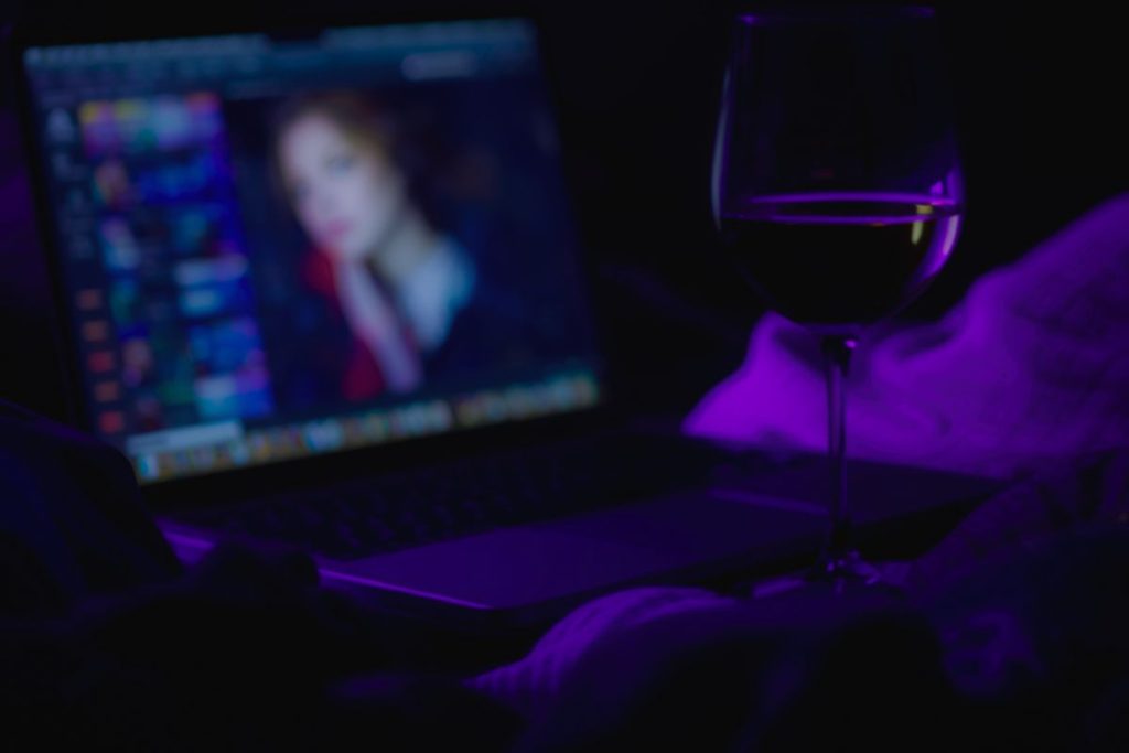 A wine glass beside a glowing laptop screen in purple light, creating an intimate late-night atmosphere.