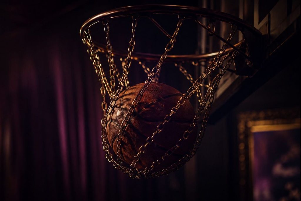 Basketball swishing through a metal chain net in moody burgundy and amber light.